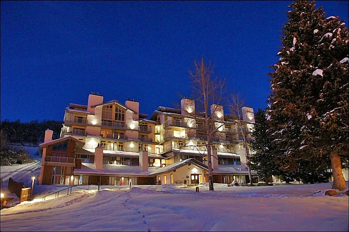 The image features an illuminated building facade framed by tall evergreen trees, with a thick layer of snow blanketing the ground. The scene exudes warmth and welcome even in winter’s chill, suggesting cozy evenings and a sense of community.