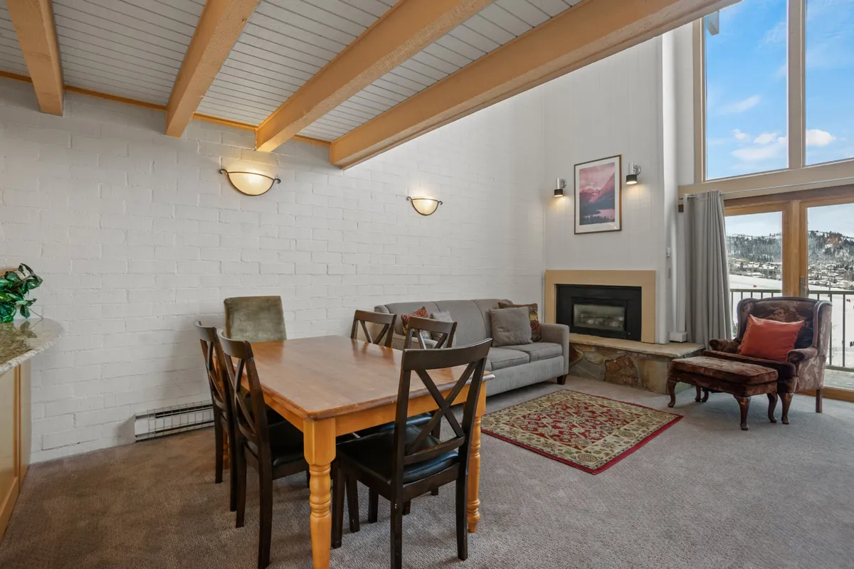 A cozy, open-concept living and dining area featuring a sofa, dining table, fireplace, and expansive windows that flood the space with natural light and mountain views. The room is spacious with vaulted ceilings, and the arrangement invites gathering for meals, conversation, or relaxing by the fire.
