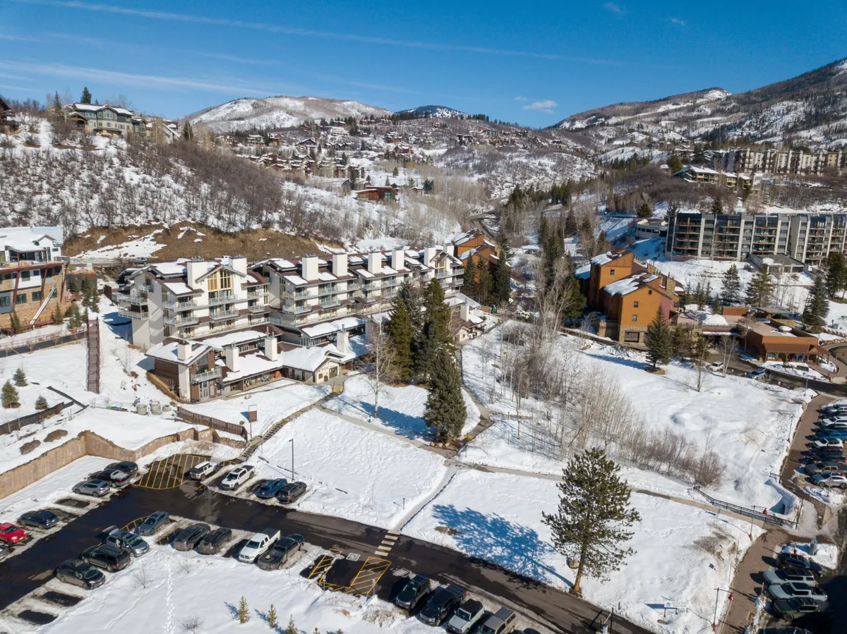 The image shows several snow-dusted buildings nestled at the base of a mountain, with a visible parking area and a line of trees under an expansive sky. The scene suggests both tranquility and adventure, with trails and ski runs just outside the door.