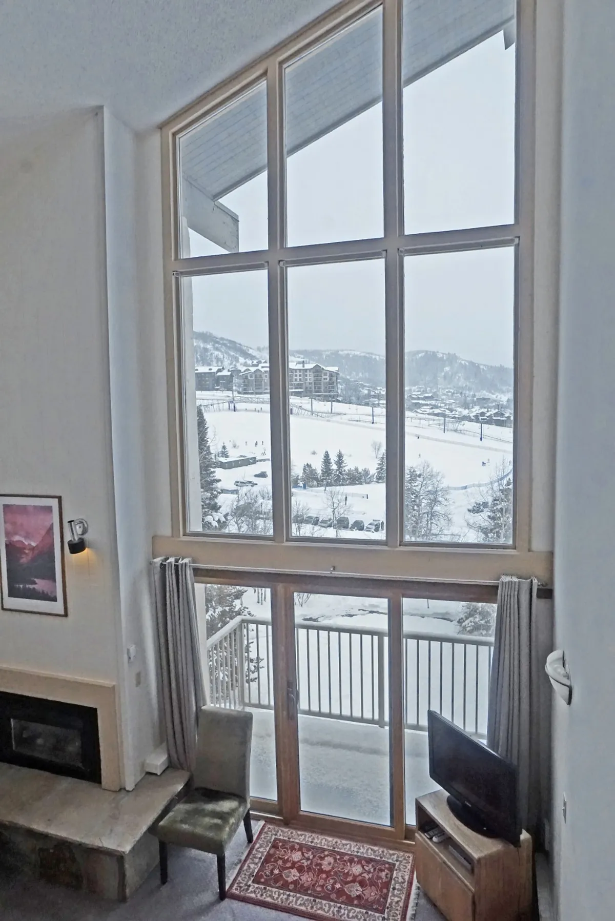 A living area with a fireplace, green chair, television, and a wall of large windows overlooking a snow-covered landscape. The balcony is visible, inviting guests to step outside and enjoy the expansive views. The space feels tranquil and ideal for gathering or quiet moments.