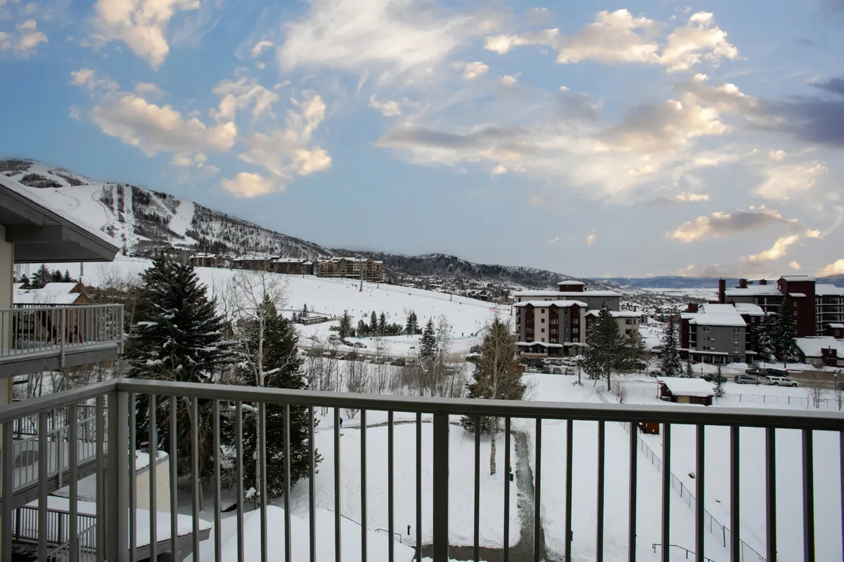 An expansive view of a snow-covered landscape, including distant ski slopes, surrounding buildings, and a cloudy sky, suggesting easy access to both nature and local amenities. The image hints at activities like skiing, enjoying hot cocoa on the balcony, and exploring nearby trails.