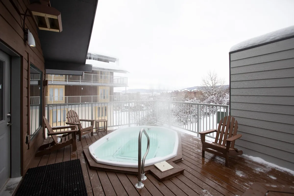 The exterior shot showcases a steaming hot tub on a wooden deck, surrounded by deep snow. Adirondack chairs invite relaxation, and the snowy landscape beyond suggests a cozy retreat after a day of skiing.
