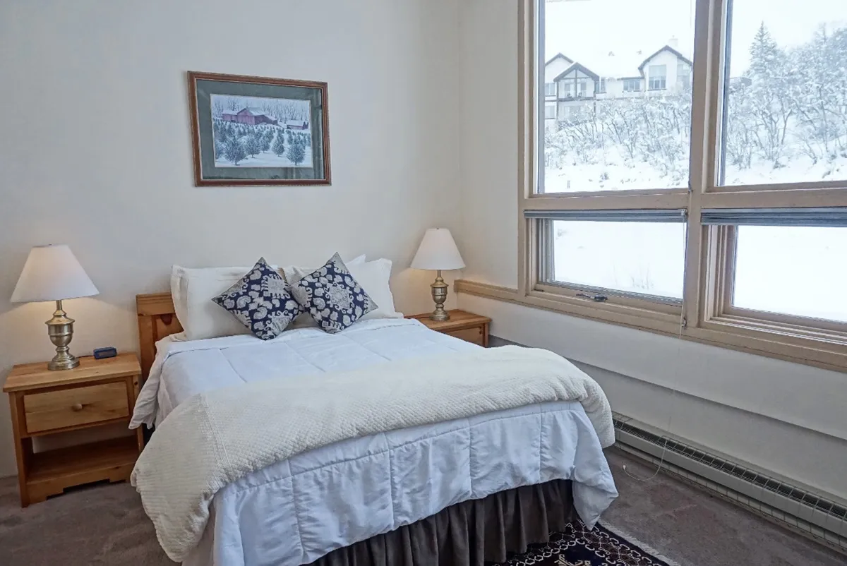 A warmly lit bedroom featuring a plush Queen bed with decorative pillows, soft lamps on either side, and a large window offering a tranquil, snowy view. Framed artwork adds a touch of personal style, while the overall ambiance is calm and inviting.