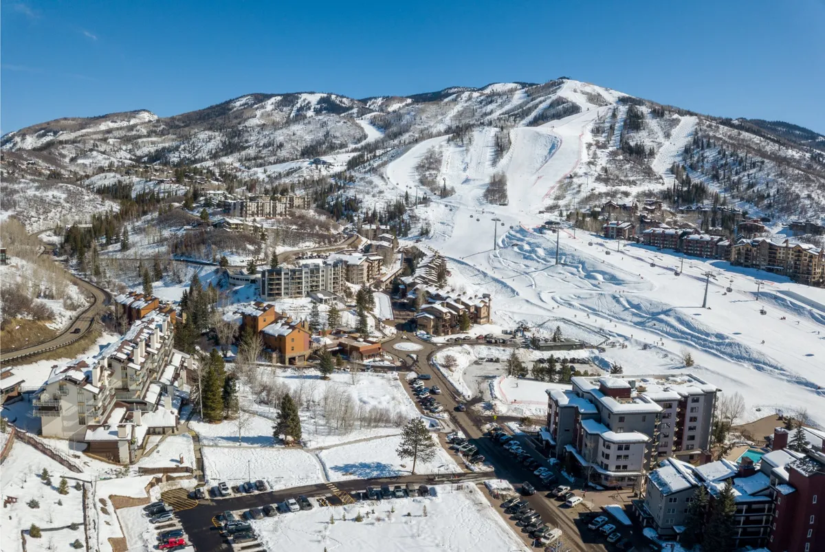 This exterior image captures the Steamboat base area with snow-covered mountains, ski slopes, residential buildings, and a visible parking area. While the image is winter-themed, it shows the proximity of outdoor recreation, the walkable layout, and the base village atmosphere central to the condo's prime location for mountain biking, hiking, and festivals in spring through fall.