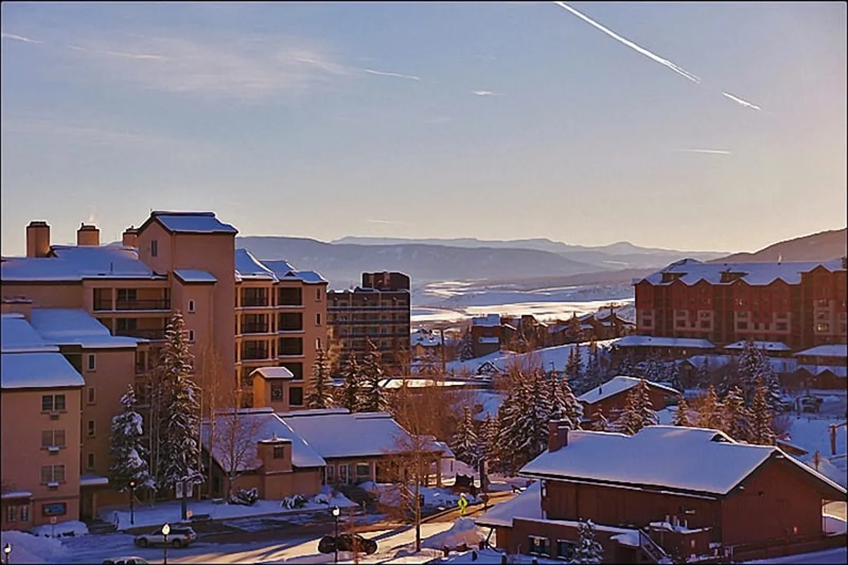 This image features snow-covered rooftops set against a backdrop of distant mountains and frosted trees, all under a glowing sunset sky. The scene evokes a tranquil, resort-like ambiance, suggesting relaxation after a day of outdoor adventure.