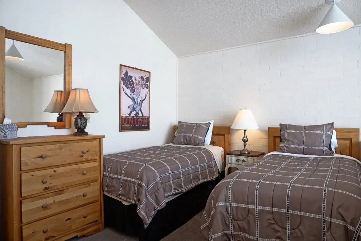 A bright loft area with two twin beds positioned parallel to each other, complemented by a dresser with a mirror, a table lamp, and a framed poster on the wall. The space is cozy yet open, ideal for kids or singles seeking independence.
