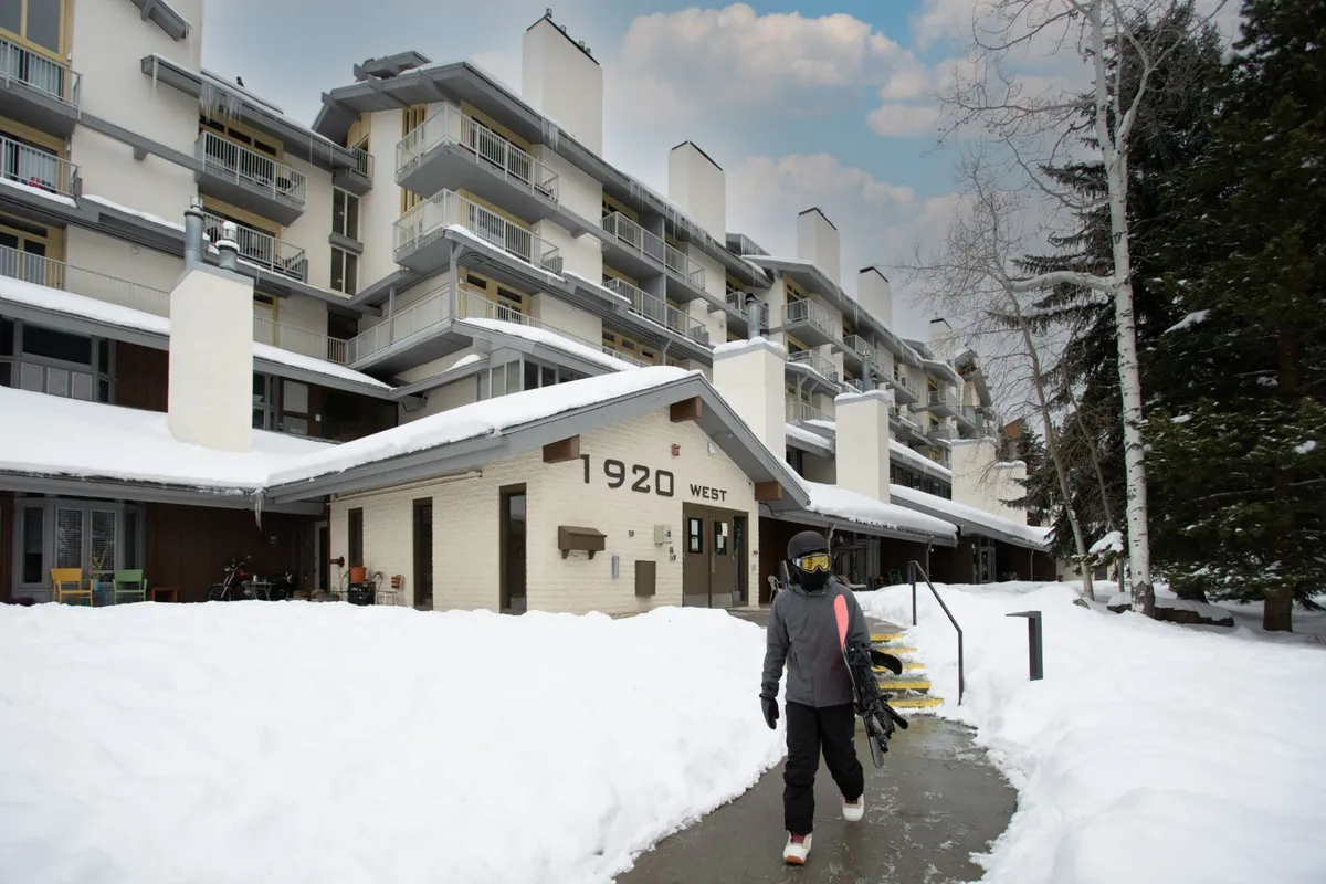 The image captures the building entrance on a snowy path, with a person carrying a snowboard, suggesting immediate proximity to the slopes. Snow blankets the landscape, and the entryway is welcoming, evoking the excitement of heading out for a day on the mountain or returning after an adventure.