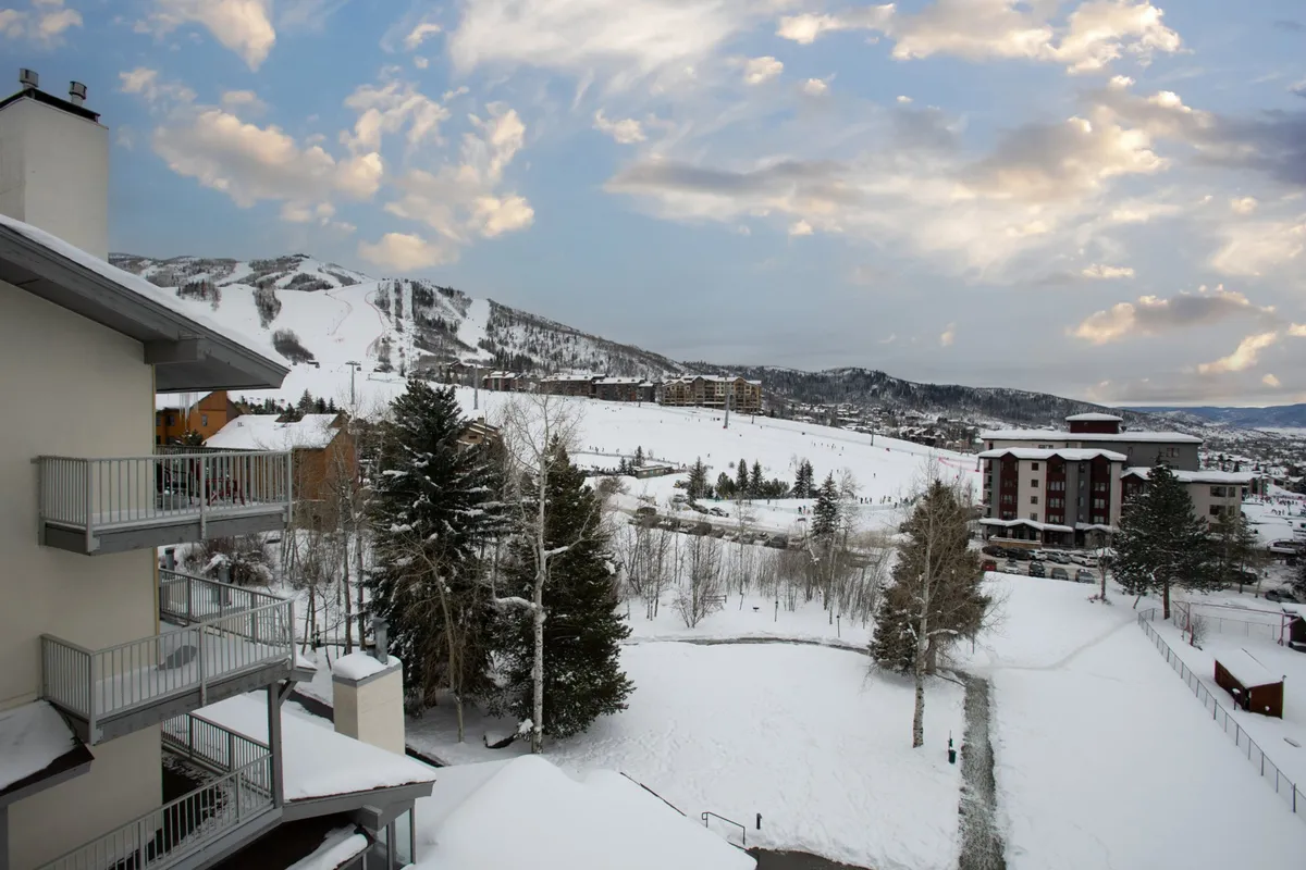 A sweeping exterior view captures snow-covered mountains rising behind ski slopes, flanked by a tree line and nearby residential buildings. The setting is serene, with clear evidence of outdoor winter activities and easy proximity to nature, reinforcing the area’s allure for extended stays and mountain living.