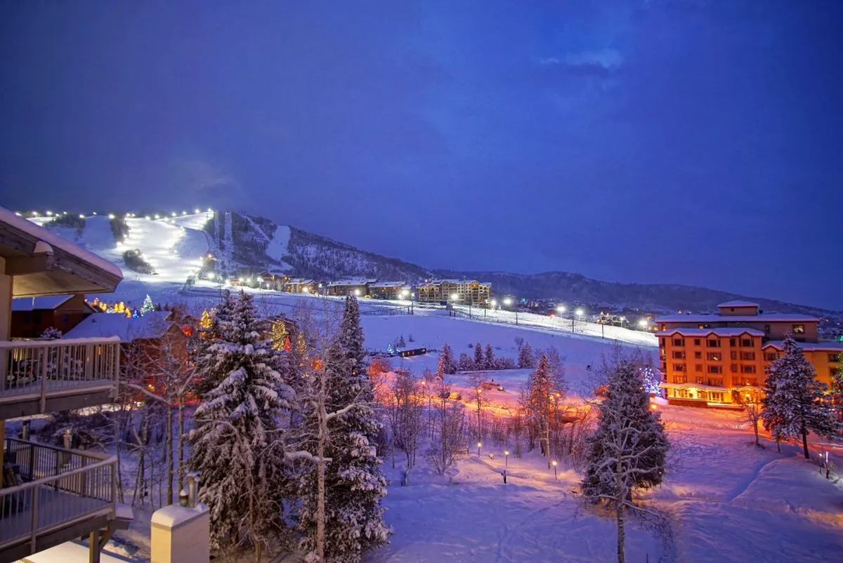 A sweeping nighttime view of snow-covered slopes illuminated for skiing, with warmly lit buildings and tall pine trees in the foreground. The scene feels magical and tranquil, capturing both action and ambiance.
