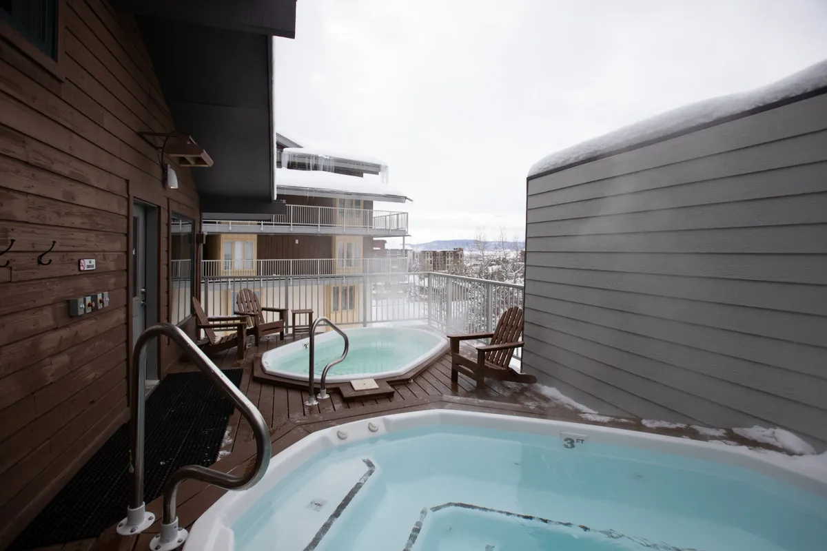 This image features outdoor hot tubs set on a wooden deck, with comfortable chairs and a balcony railing. The snowy landscape visible in the background underscores the resort-style relaxation possible after a day in the mountains.